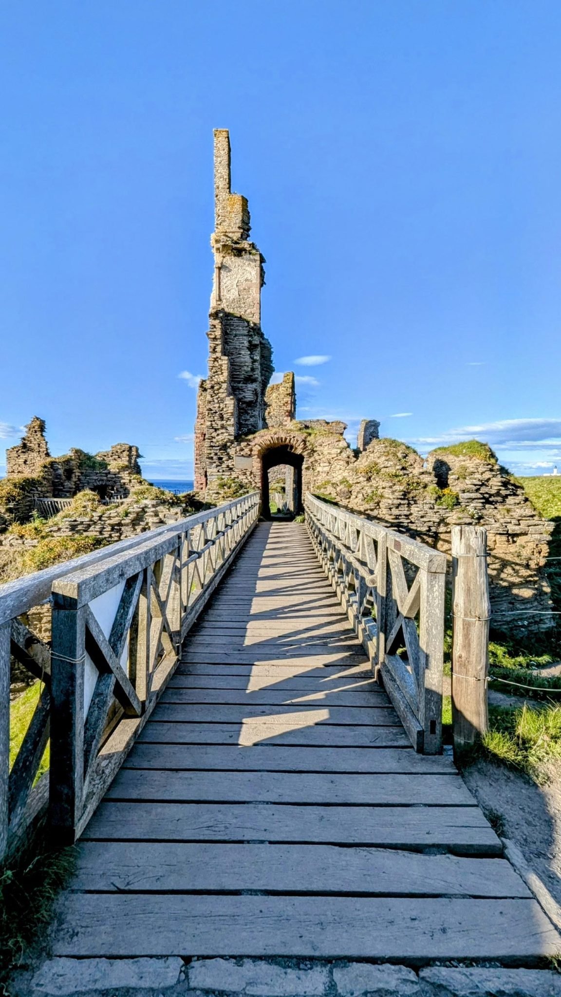 A wooden footbridge leading across a deep rock gully to the arched gateway of Castle Sinclair Girnigoe, its tall splinter of ruined tower against a blue sky, Noss Head lighthouse visible in the distance.