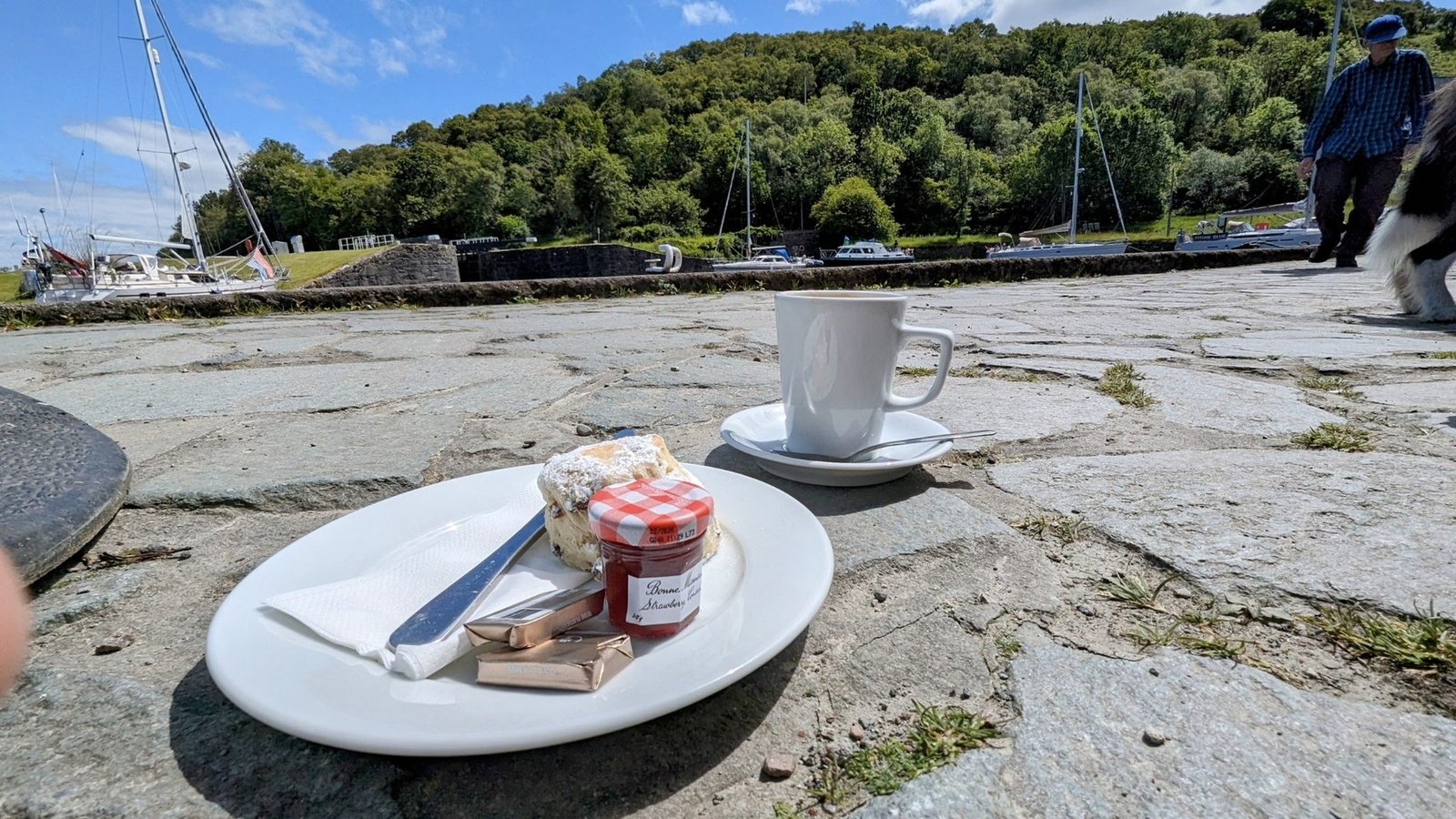 Coffee and a slice of cake on a plate on the rocks at Crinan, boats and sea behind.