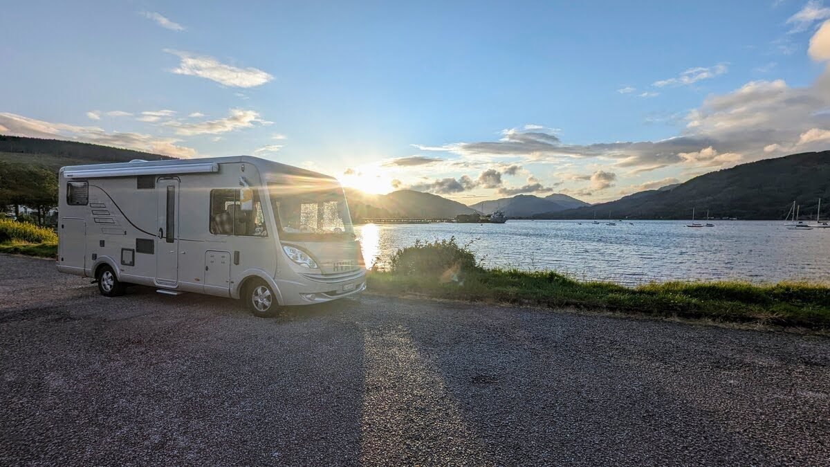 A motorhome parked at a grassy shoreline pull-in near Dunoon at sunset, the sun setting over the hills across the Firth of Clyde.