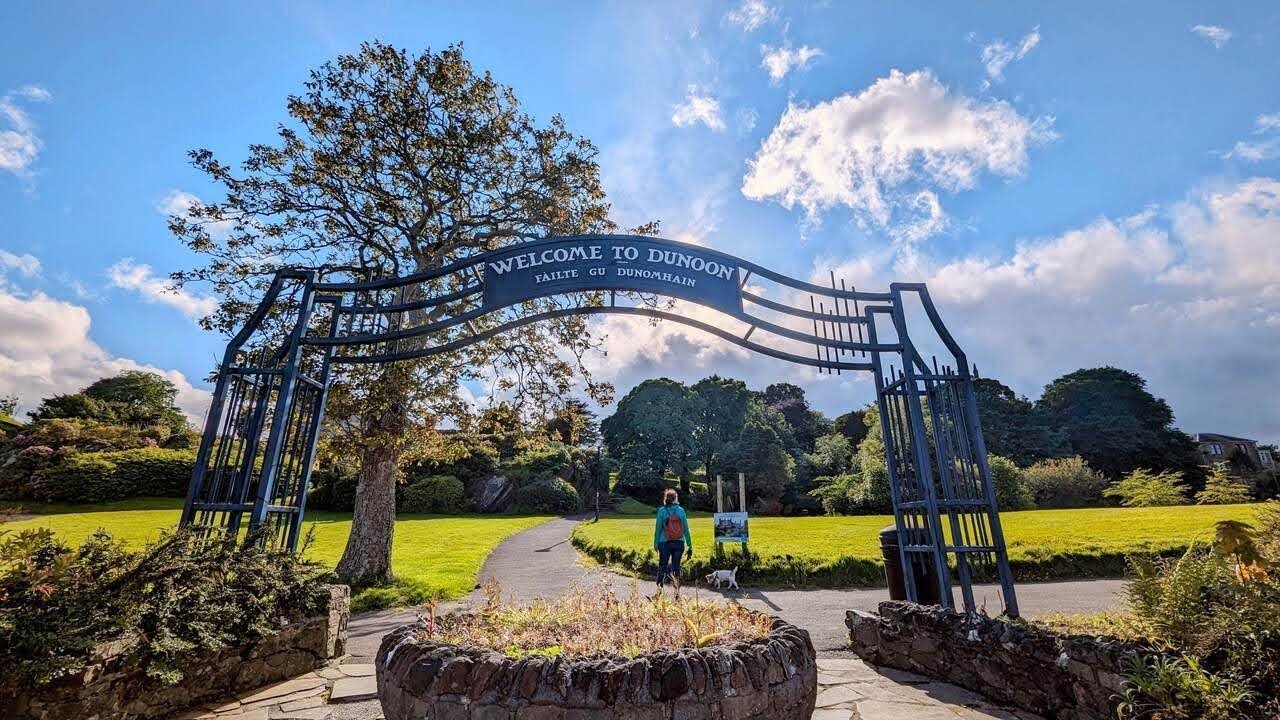 The ornate metal 'Welcome to Dunoon' arch over a path in a seafront park, hills beyond.