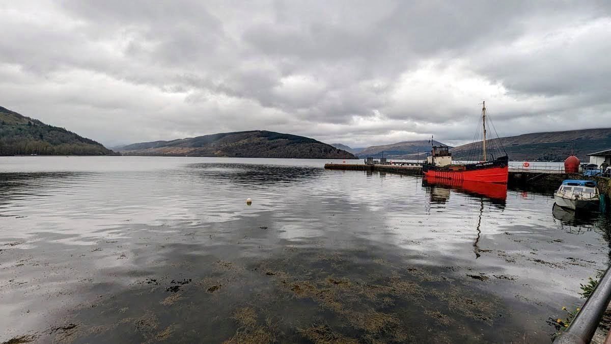 A red fishing boat moored at the Inveraray pier on Loch Fyne under a grey sky.