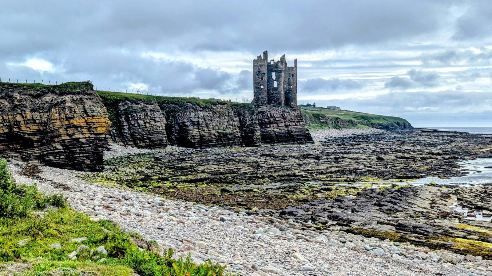 Keiss Castle ruin on its cliff seen from a pebble-and-flagstone beach at low tide, layered rock strata in the cliff face.