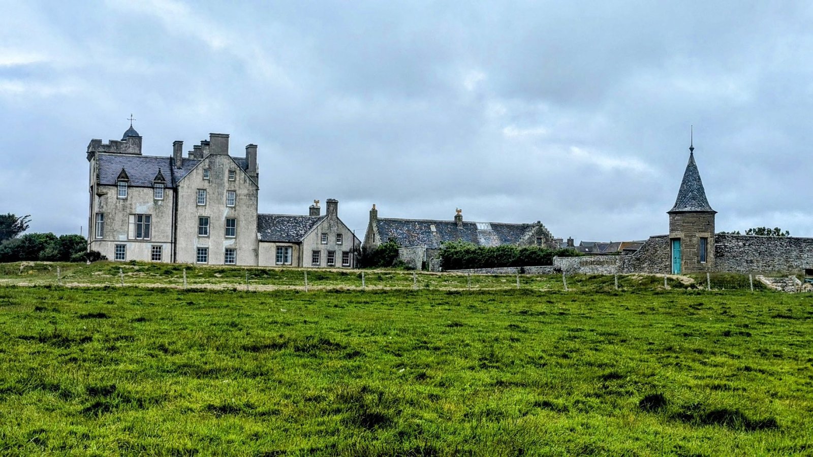The grey Scottish Baronial mansion of 'new' Keiss Castle with crenellated tower, chimneys and a pointed-roof turret, behind a green field.