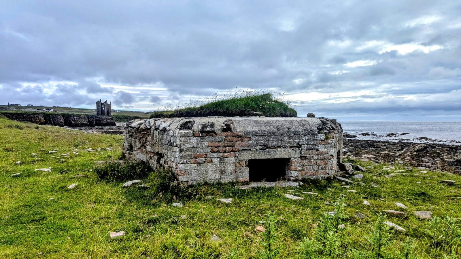 A WWII brick pillbox in the foreground on grass, with the ruined tower of Keiss Castle on the clifftop in the distance.