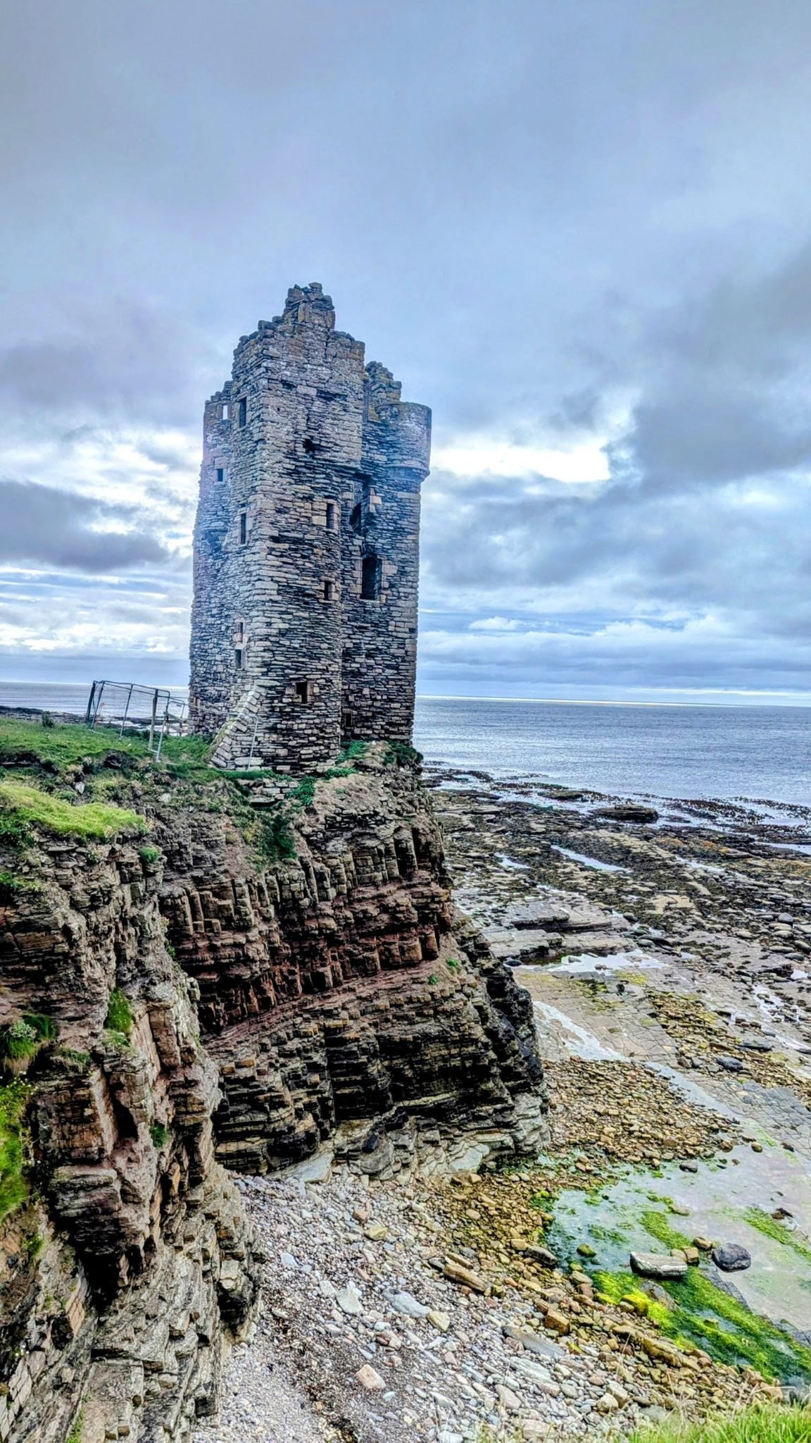 The tall ruined stone tower of Keiss Castle standing on the edge of a layered sandstone sea cliff above a low-tide rock platform, Caithness.