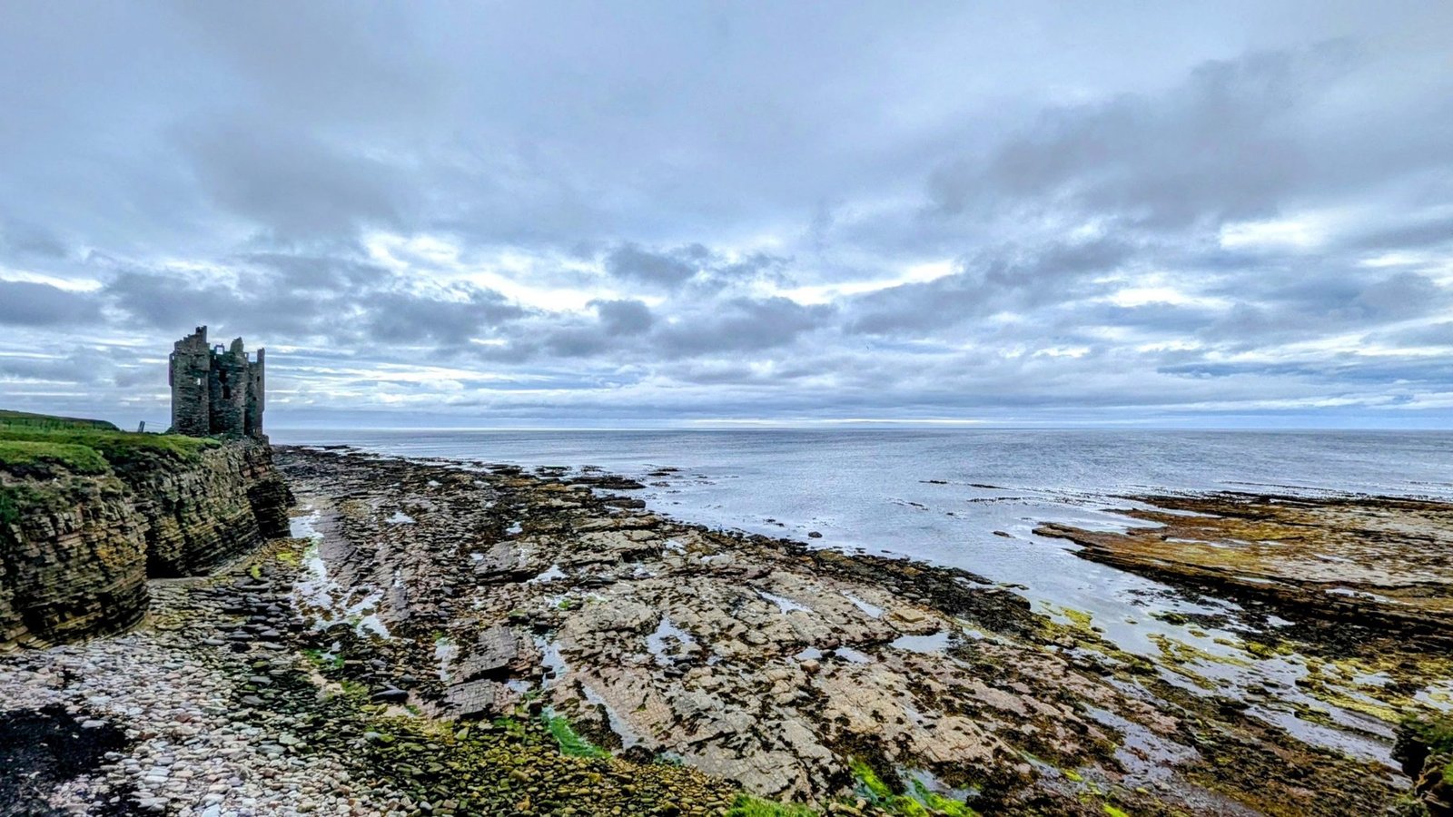 Wide dawn view of the Caithness coast at Keiss with the castle ruin on the left headland and rock platforms stretching into a calm sea.