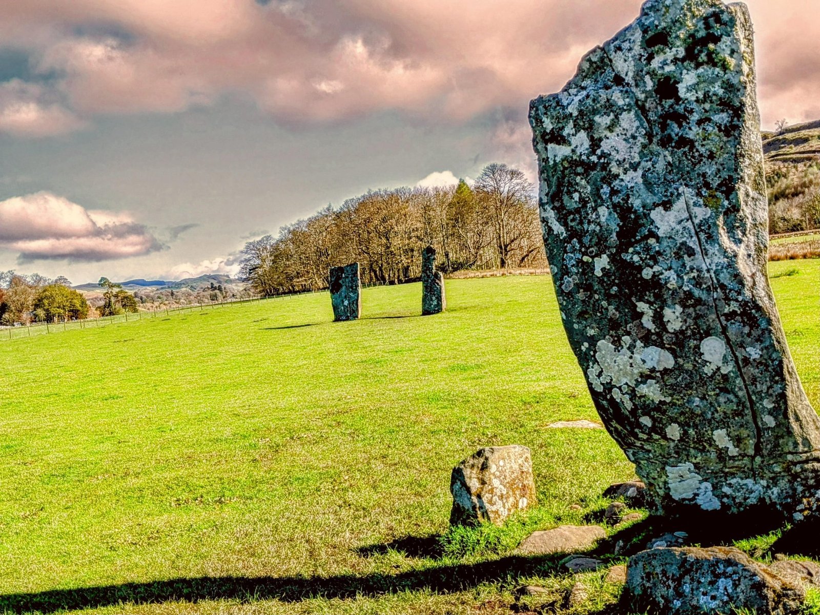 A tall, lichen-covered prehistoric standing stone leaning against a dramatic sky in a green field at Nether Largie, Kilmartin Glen.