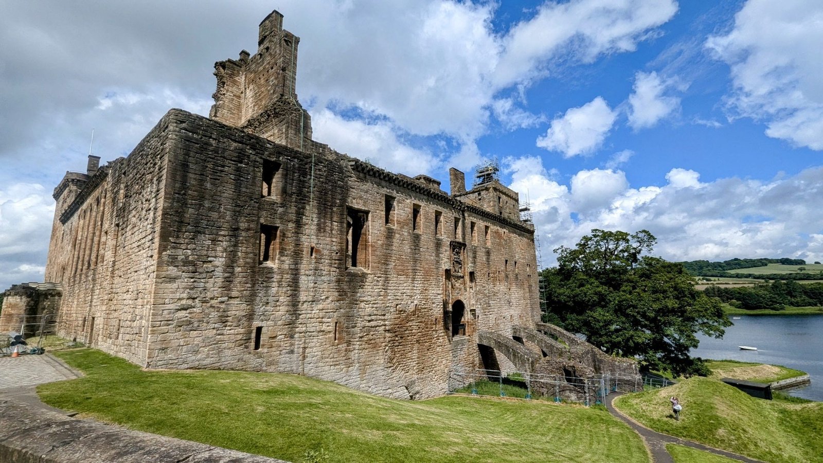 The tall ruined stone walls of Linlithgow Palace above green lawns beside Linlithgow Loch under a blue sky.