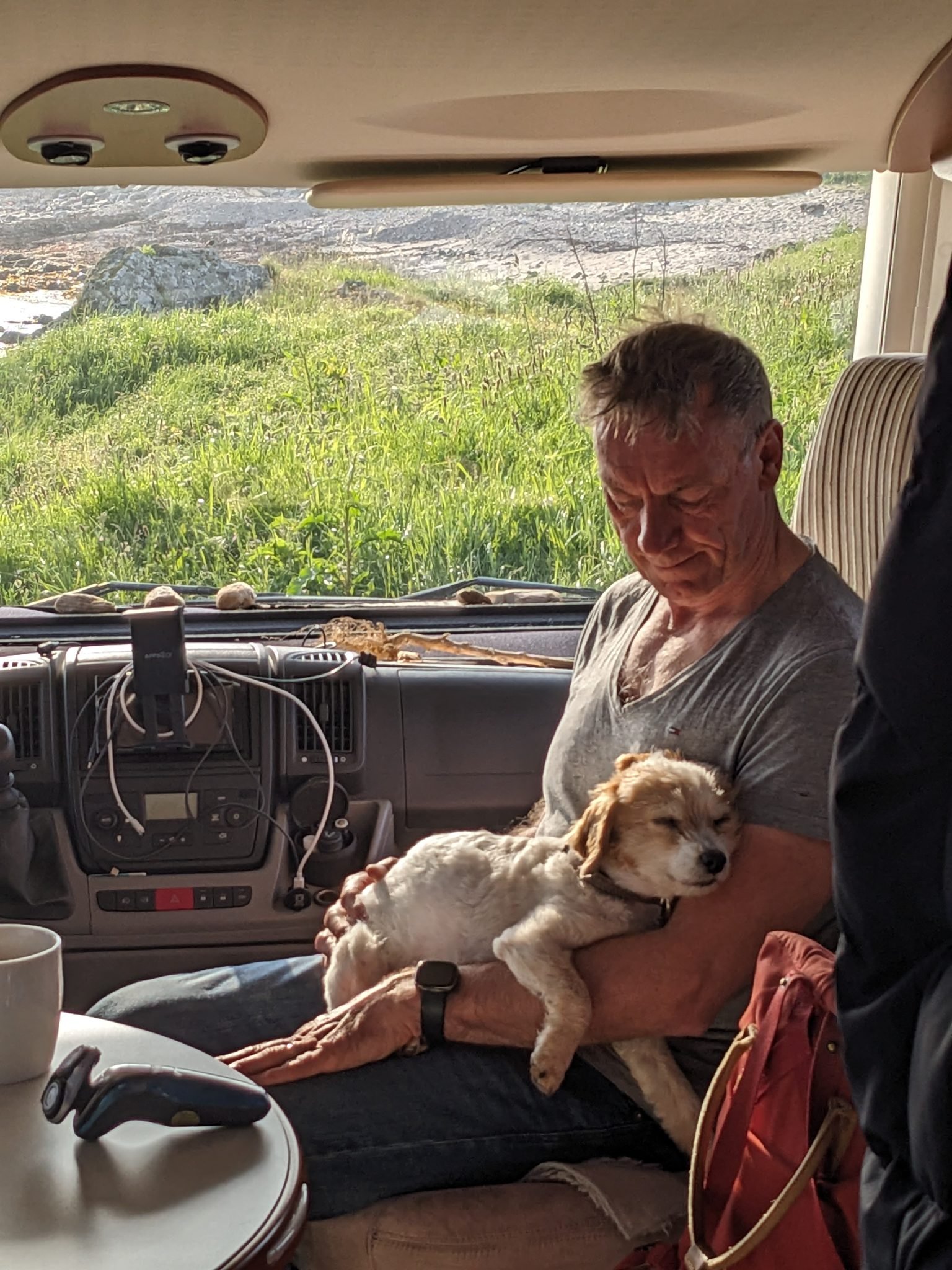 Inside the motorhome at Machrihanish in the morning — a man relaxing with a small dog on his lap and coffee, sea view through the window.