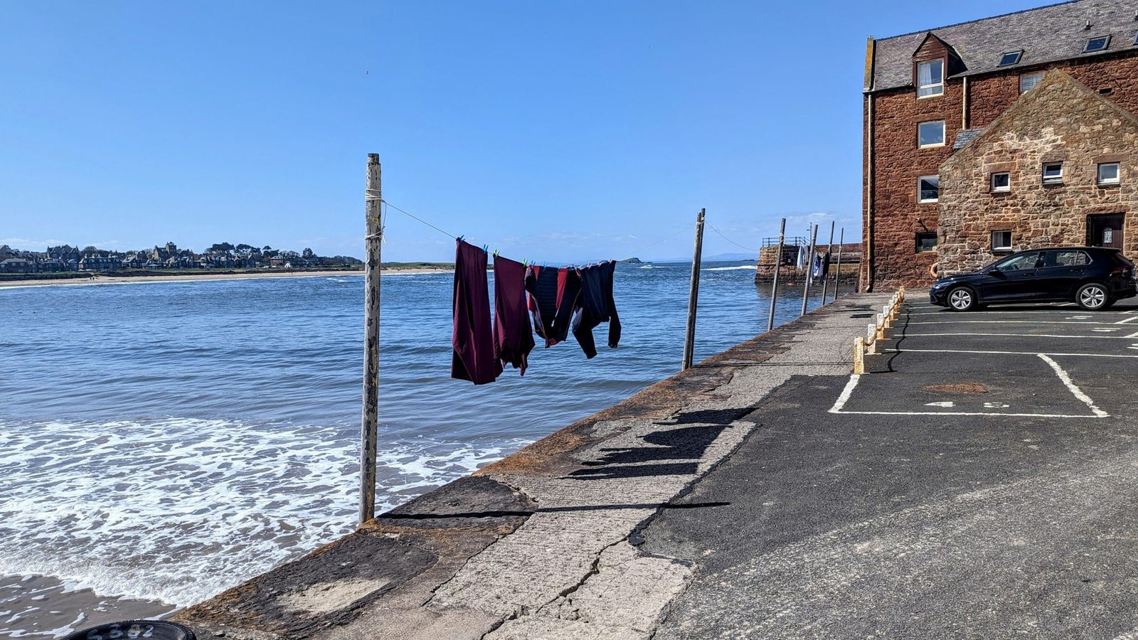 Swimwear and wetsuits drying on a line strung along the harbour wall at North Berwick, West Bay beach and the town beyond under a blue sky.