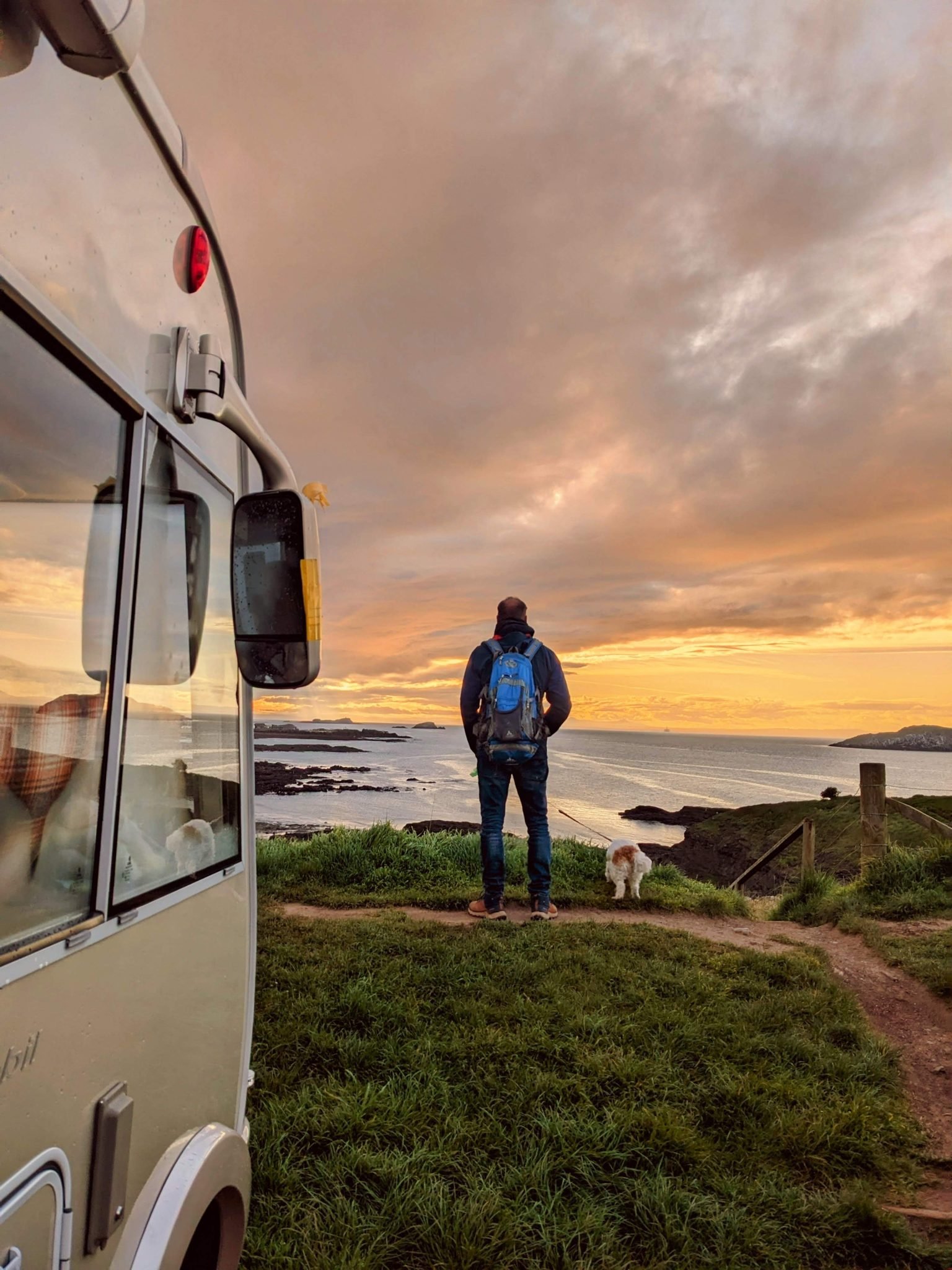 A Hymer motorhome parked on a grassy clifftop at North Berwick at sunset, with a person and dog looking out to sea.