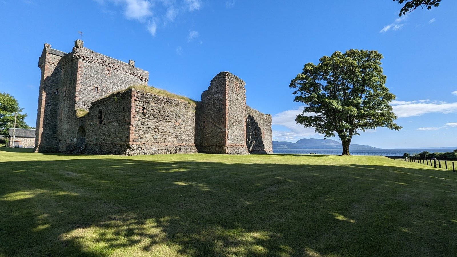 The stone curtain walls and tower house of Skipness Castle on green grass, a lone tree and the mountains of Arran across Kilbrannan Sound behind.