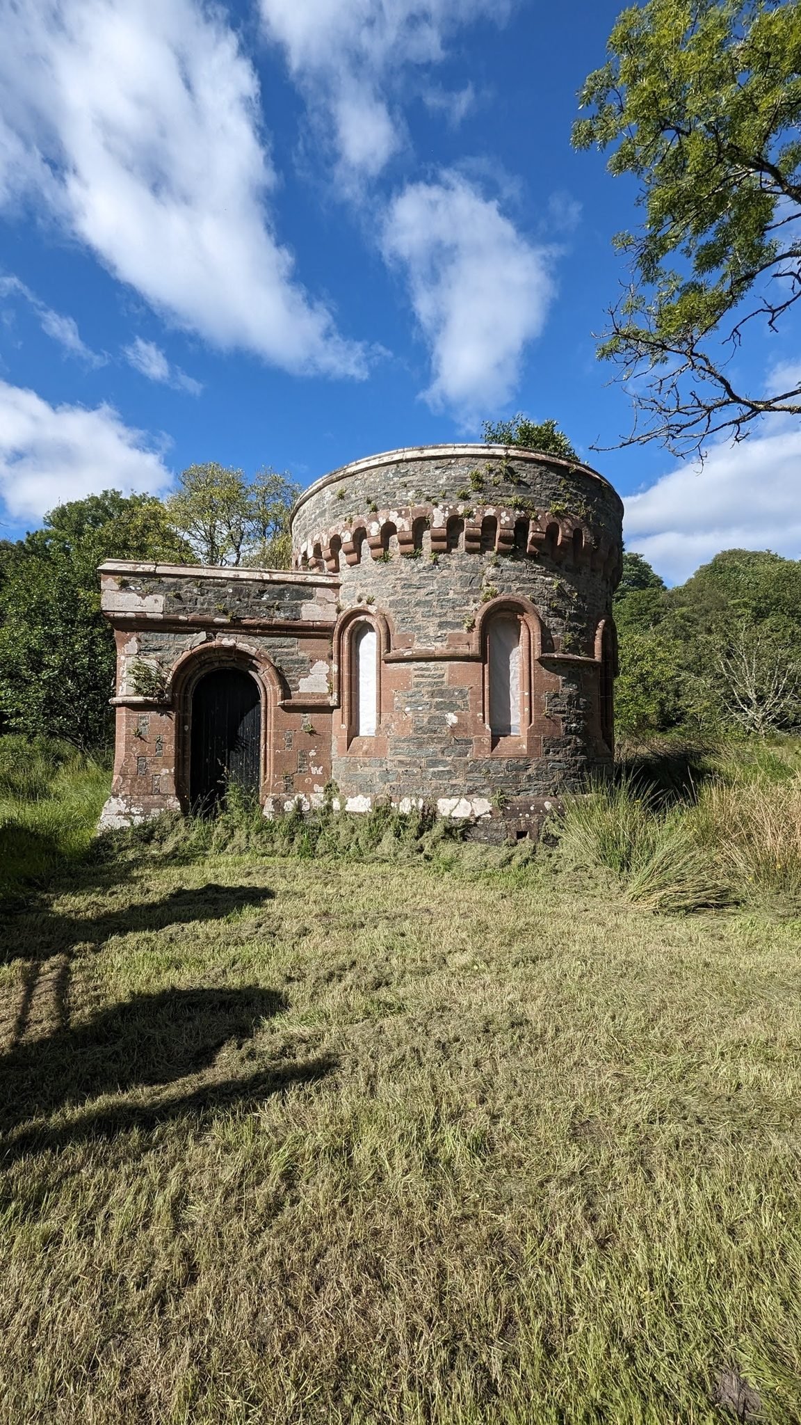 An ornate red-sandstone castellated round tower folly standing in long grass among trees at Skipness.