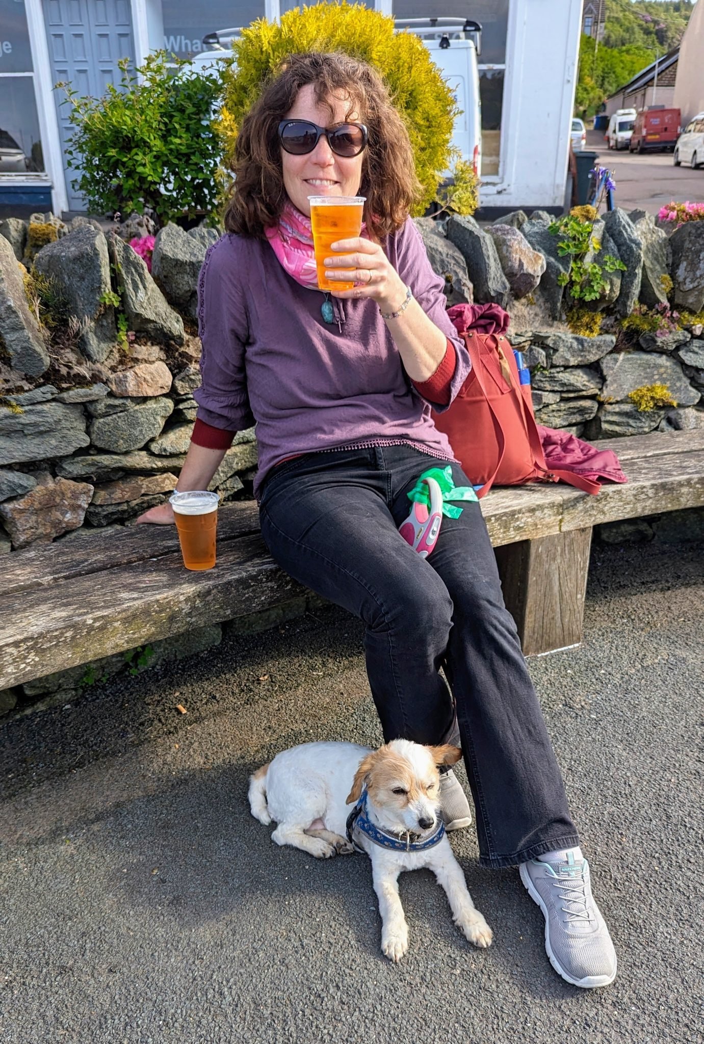 A woman with a beer sits on a harbour bench in Tarbert, a small dog at her feet.