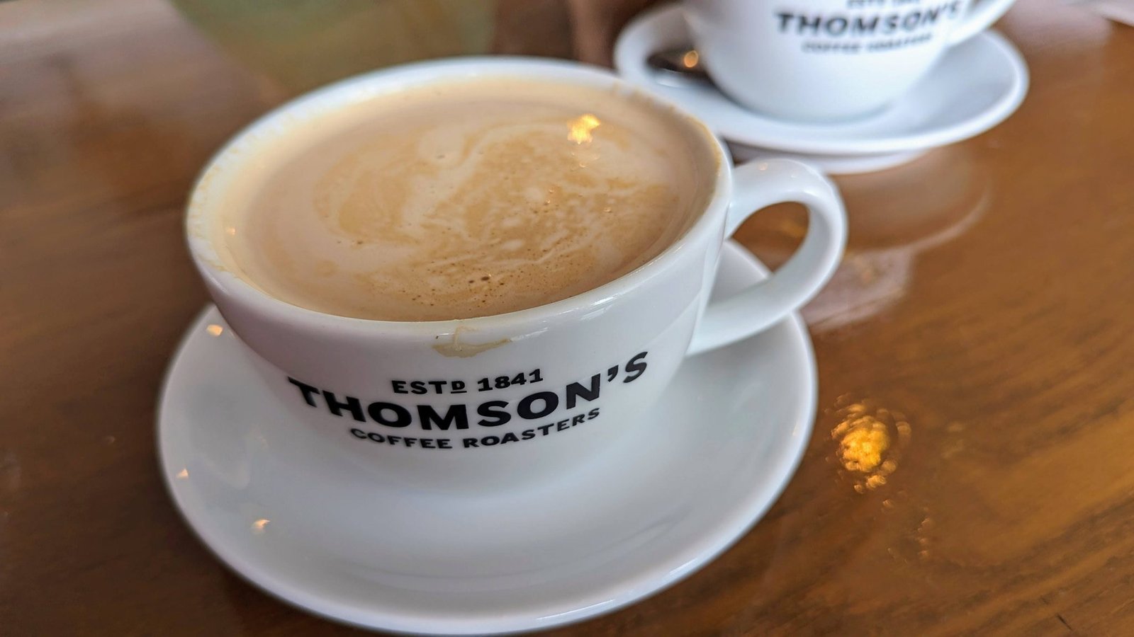 Two white cups of coffee marked 'Thomson's Coffee Roasters, est. 1841' on a wooden café table.