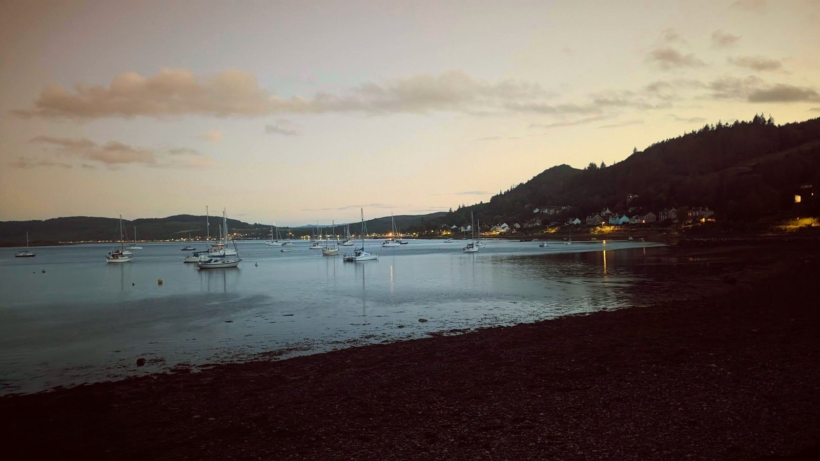 Moored yachts silhouetted on the Kyles of Bute at dusk, village lights beginning to show.