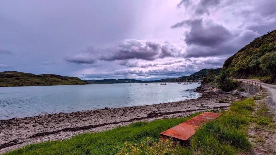 The quiet single-track shore road along the Kyles of Bute near Tighnabruaich under a moody sky, water and hills beyond.