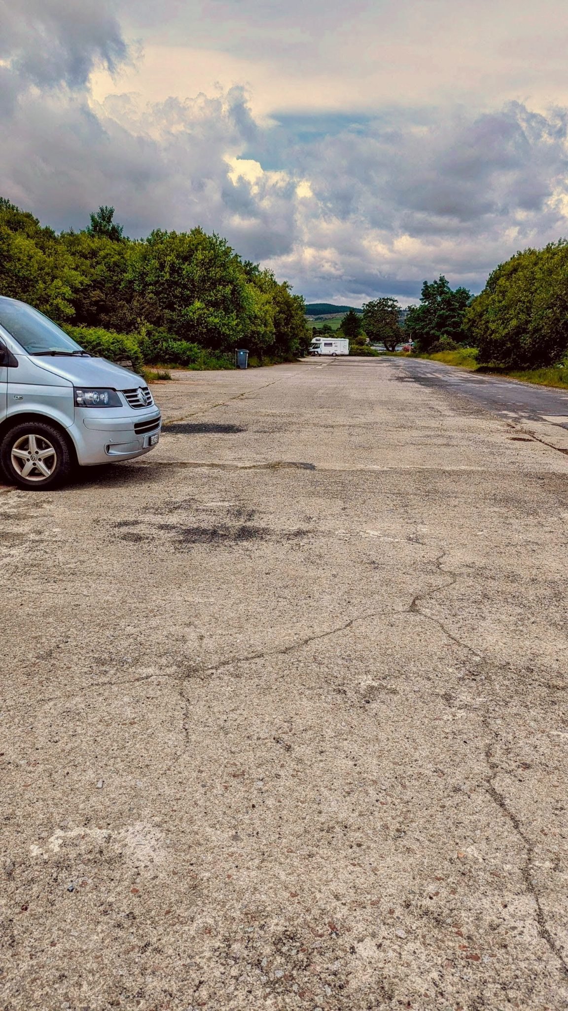 A VW T5 campervan parked on a large flat tarmac area near Tighnabruaich, trees and hills behind.