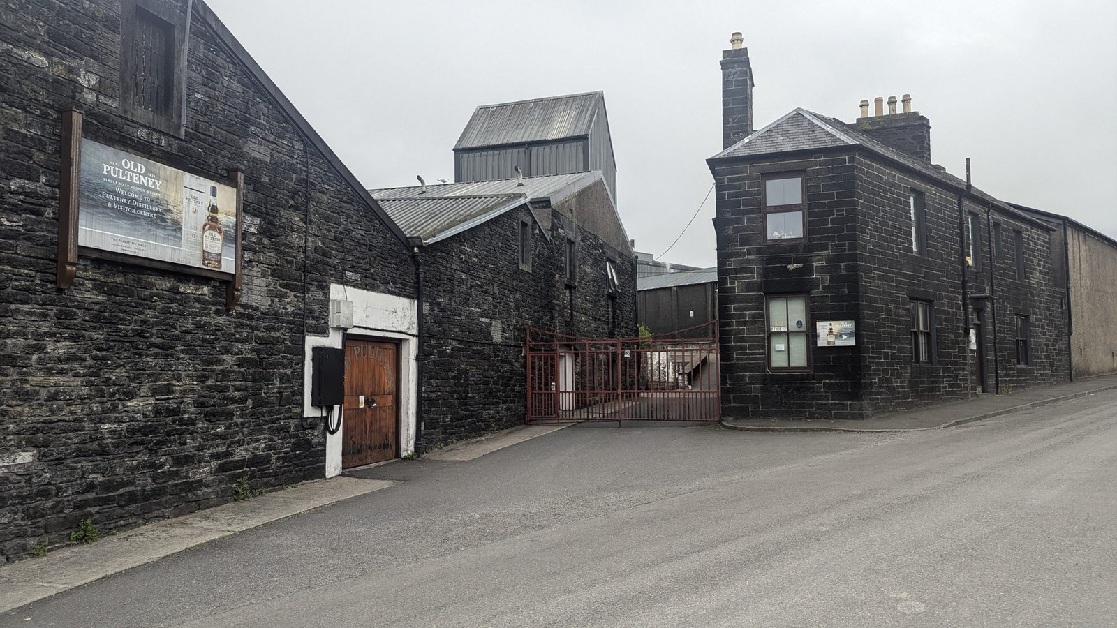The dark Caithness-flagstone buildings of Old Pulteney distillery in Wick on a misty morning, with a metal malt-kiln roof and the Old Pulteney welcome sign.