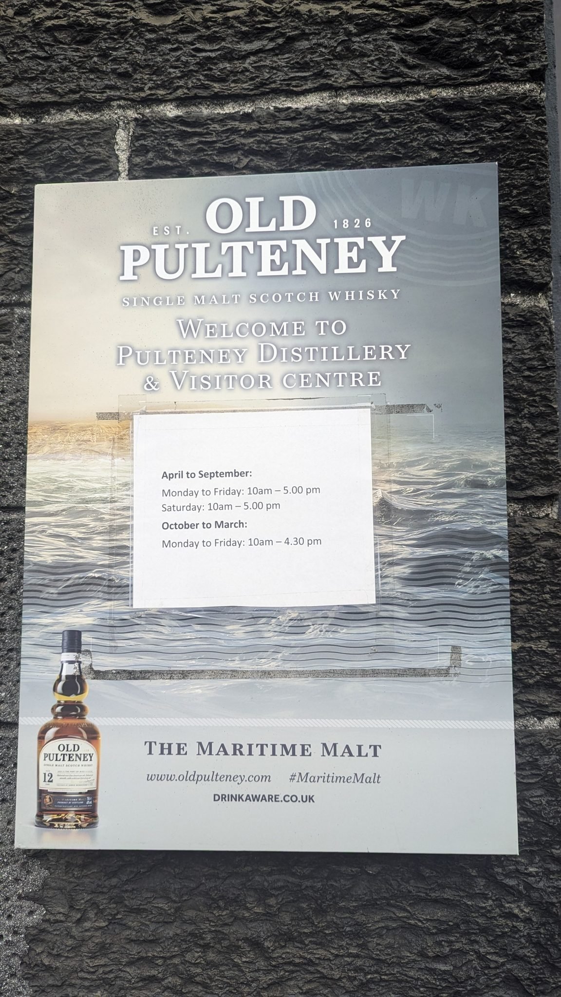 Old Pulteney distillery welcome sign on a flagstone wall listing visitor-centre opening hours.