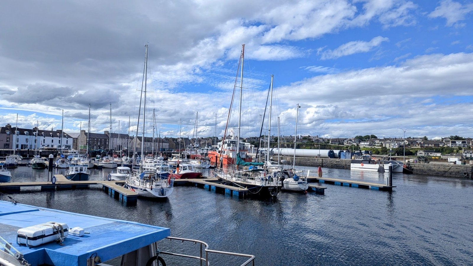 Yachts moored at pontoons in Wick marina with the stone terraces of Pulteneytown behind and offshore wind-turbine tower sections stacked on the quay.
