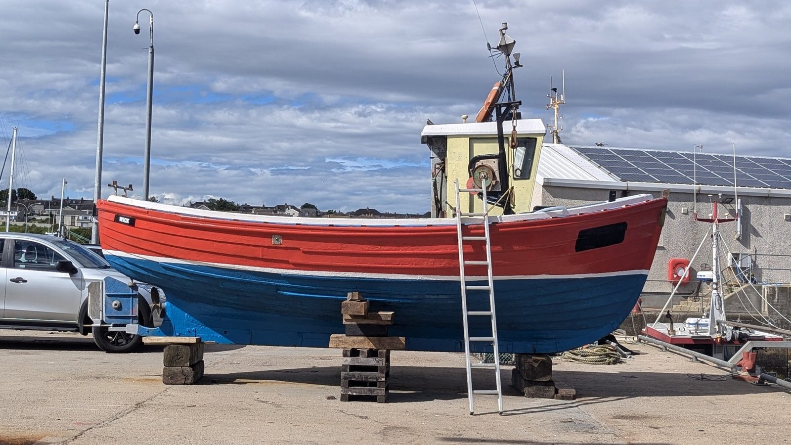 A traditional clinker-built wooden fishing boat painted red, white and blue, up on blocks on the quay with 'WICK' on the bow and a ladder against it.
