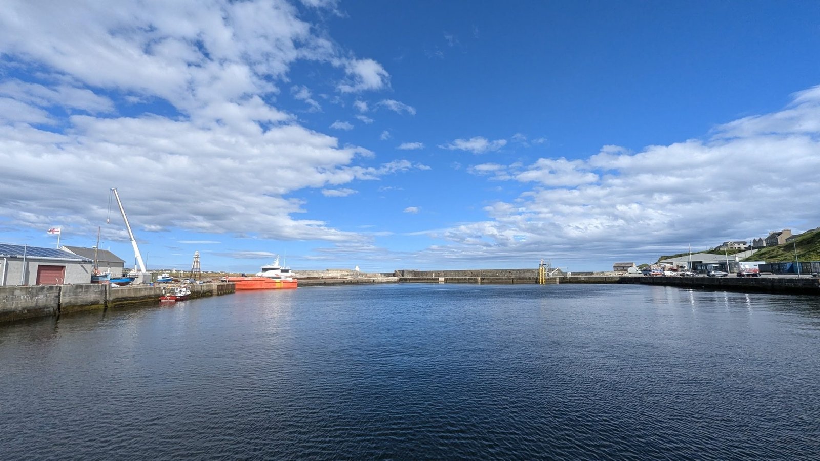 Wick's outer harbour with stone quay walls, a crane, an orange offshore-industry vessel and harbour buildings under a bright sky.